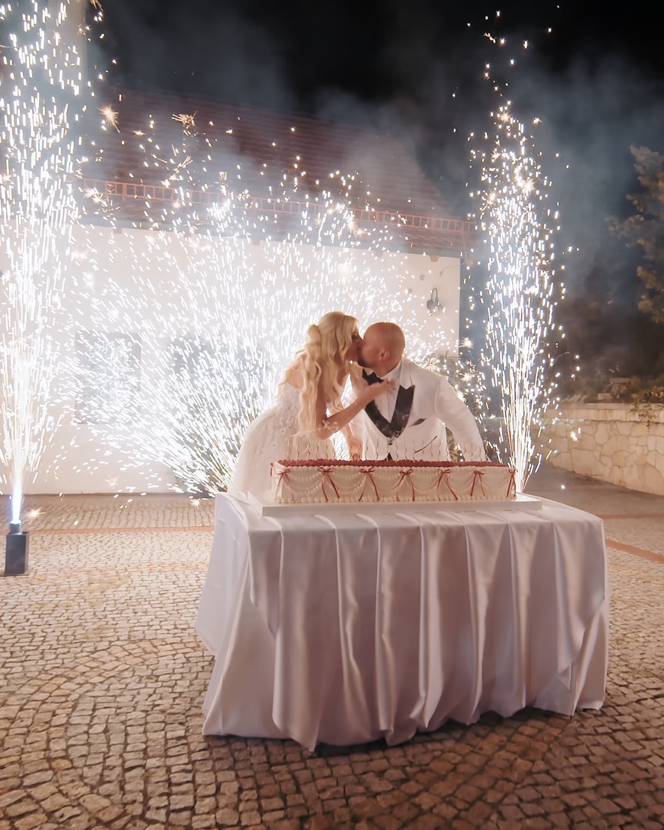 Para młoda całuje się przy torcie na dziedzińcu nocą, za nimi fontanny iskier